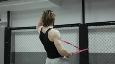 Siyah kollu tişörtlü sporcunun, kol kaslarıyla çalışmak için spor aletlerine verilen dayanıklılık bandını kullanışının arka planı. Motive olmuş erkek, içeride ısınmak için süspansiyon egzersizleri yapıyor..