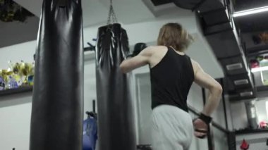 Siyah kum torbalarının önüne atlarken bacağıyla tekmeleyen konsantre beyaz erkek. Erkek sporcu nefsi müdafaa teknikleri üzerinde çalışıyor ve parlak paspas üzerinde dayanıklılık çalışması yapıyor..