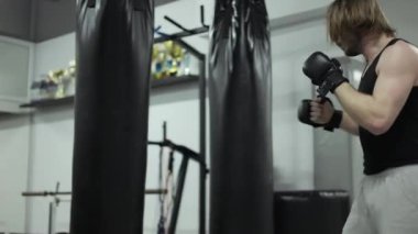 Siyah eldiven giyerken siyah kum torbasıyla el ve bacak alıştırması yapan beyaz bir adam. Rahat kıyafetler içindeki güçlü adam spor salonunda tek başına antrenman yapıyor ve dövüşten önce egzersiz yapıyor..