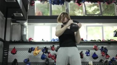 Uzun saçlı genç adam elleriyle yuvarlak hareketler yapıyor ve kettlebell kullanarak egzersiz yapıyor. Çalışkan sporcu rekabet için vücut kaslarını güçlendirmeye odaklanıyor.