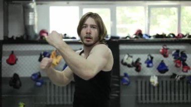 Kafkasyalı, uzun saçlı, spor salonunda dövüş sanatları antrenmanı yapan, duvarlarında renkli boks eldivenleri olan bir adam. Kendini savunma eğitmeni. Gri duvarlı odada kol ve bacakları kullanarak teknikler gösteriyor..