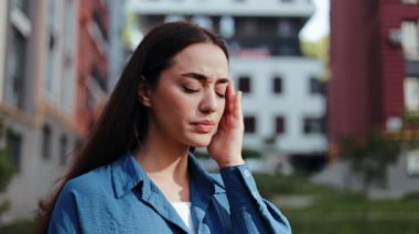 Uzun kahverengi saçlı kadın baş ağrısı hisseder ve elleri sokakta dururken şakaklarını ovuşturur.