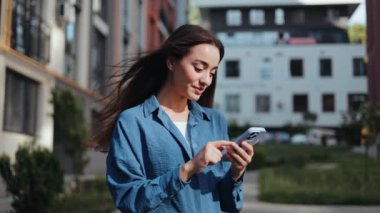 Güneşli bir günde dışarıda yürüyüp akıllı telefon kullanan mutlu genç bir kadın.
