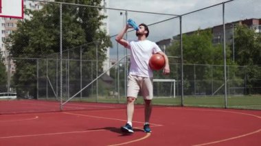 Yakışıklı adam mavi plastik şişeden bir yudum su yapıyor ve elinde basketbol topu tutuyor. Beyaz tişört giymiş aktif bir erkek açık havada su dengesini sağlıyor. Sıvı kavramı.