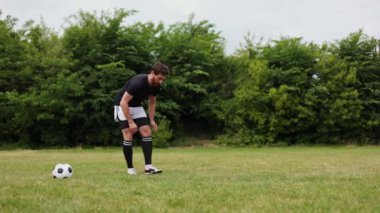 Kafkasyalı erkek çimenlik alanda esnemeye hazır, futbol topu da yakında. Siyah şortlu genç sporcu ve siyah kollu beyaz gömlekli açık hava egzersizleri yapıyor..