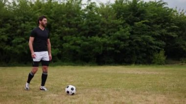 Aktif adam siyah bluz ve beyaz şort giyerek futbol topunu çimenli sahada tekmelerken elini kaldırdı. Ağaçların ve açık gökyüzünün arkaplanı. Futbolun açık havada oynandığı an.