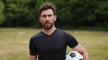 Kendine güvenen beyaz bir oyuncunun portresi spor üniforması giymiş gür ağaçların ve çalıların üzerinde geniş çayırlarda geziniyor. Futbol topu tutan yakışıklı genç adam ve yana bakıyor..