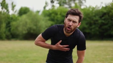 Siyah üniformalı bitkin bir adam göğsünü eliyle tutuyor ve doğa arka planında tek başına dikilirken nefes almakta zorlanıyor. Stresli sporcu dışarıda koştuktan sonra antrenmanı bırakıp mücadele ediyor..
