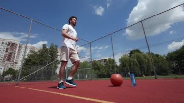 Proaktif beyaz sporcu bacağını kaldırıyor ve dirseğiyle dizine dokunuyor. Yanında basketbol topu ve bir şişe su da var. Açık mavi gökyüzü altında geniş bir basketbol sahası. Zindelik kavramı.