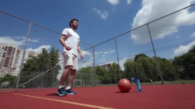 Aktif beyaz sporcu bacağını kaldırıyor ve dirseğiyle basketbol ve su şişesiyle dizine dokunuyor. Basketbol sahası ve açık mavi gökyüzü. Egzersiz, eğitim, hobi ve spor kavramı.