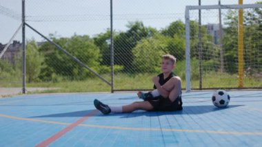 Spor üniformalı şirin bir çocuk. Bacağını dizine kadar eğiyor ve futbol malzemeleriyle kol kola girip ayağını çeviriyor. Burkulma ve kramplardan kaçınmak için hareket eden kaygısız bir çocuk..