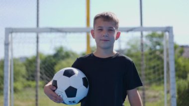 Spor sahasının arka planında futbol topunu tutarken kameraya doğru giden kendine güvenen sarışın çocuğun portresi. Gururlu çocuk gündüz antrenmanından sonra stadyumu terk ediyor..