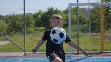 Siyah elbiseli şirin bir çocuk tek renkli futbol topuna bakarken dizleri ve ayakları boş futbol kapılarının arkasında tekmeliyor. Kaygısız çocuk spor malzemeleriyle temiz hava alıyor..