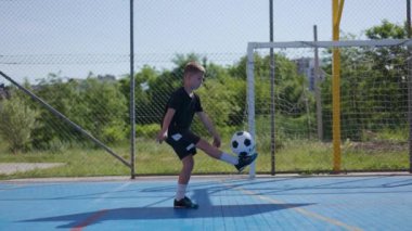 Sarışın çocuk futbol sahasının ortasında duruyor ve sağ ve sol bacaklarının dizleri ve ayaklarıyla topa vuruyor. Hevesli genç spor için içeri giriyor ve arkadaşlarını bekliyor ve oyun oynuyor..
