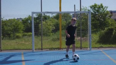 Futbolcu çocuk formalı ve şortlu. Kapıdan sahaya koşuyor. Üzerinde mavi yüzey ve işaretler var. Çalışkan genç rakiplerinin hareket ve kayma hızını kullanıyor..