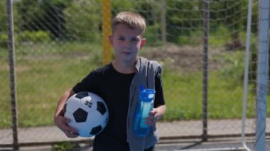 Okul müsabakaları sırasında başarılı bir maçtan sonra beyaz ağla futbol kapısından çıkan sarışının ön görüntüsü. Sıcak bir günde ferahlamak için top ve içme suyu taşıyan pozitif kaleci..