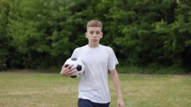 Kendine güvenen beyaz bir çocuğun çimlerin üzerinde yürüdüğünü açık ormanın yakınında ve elinde futbol topu tutarken. Beyaz tişört giymiş yetenekli bir genç futbol antrenmanına hazırlanıyor..