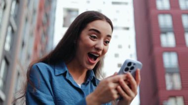 20 'li yaşlardaki bir kadın, şehir caddesinde dikilirken akıllı telefonuna heyecan verici bir tepki veriyor.