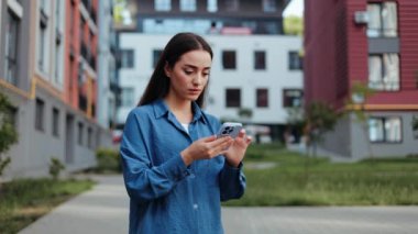 Şehir kaldırımında mavi gömlekli bir kadın akıllı telefonunu sosyal medya için kullanıyor ve endişeli görünüyor.