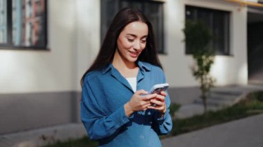 Millennial Woman in Blue Shirt Kentin dışında akıllı telefon kullanıyor.