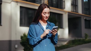 Gülümseyen kadın telefonuyla sosyal medyada geziniyor, şehir ortamında güneşli bir günün tadını çıkarıyor..