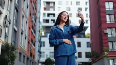Mutlu genç bir kadın şehir caddesinde telefonuyla görüntülü konuşma yapıyor. Gülümsüyor ve kameraya el sallıyor..