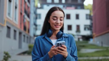 Genç ve mutlu bir kadın akıllı telefonunu kullanarak ve kulaklıkla müzik dinleyerek dışarı çıkıyor..