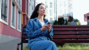 Bir bankta otururken akıllı telefon kullanan mutlu genç bir kadın..