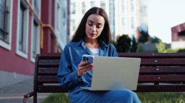 Bilgisayarında uzaktan çalışan bir iş kadını telefonunu kontrol ederken parkta bir bankta oturuyor..