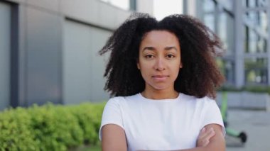 Çalı saçlı, ciddi bir kadın modern bölgede yürümek için sade beyaz tişört giyiyor. Kamera karşısında el ele tutuşan gururlu bir kadın ofis binasıyla ilgili projeler üzerinde düşünüyor..