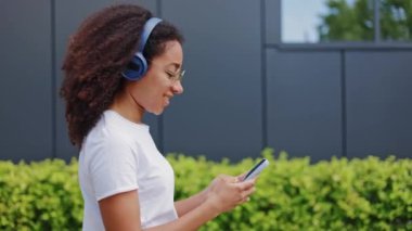Siyah modern duvarların ve yeşil çalıların yanında yürüyen ve kablosuz kulaklıkla müzik dinleyen Afro-Amerikan kadın. Güzel genç bayan dışarıda akıllı telefonlara bakıyor..