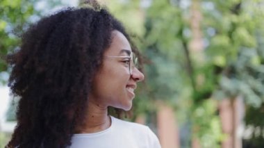 Yakından ve yandan afro-amerikan kadınını dışarıda dikilirken ve açık havada gözlük takarken görüyorum. Sokakta ağaçlarla kaplı beyaz tişört giymiş neşeli genç bir kadın..