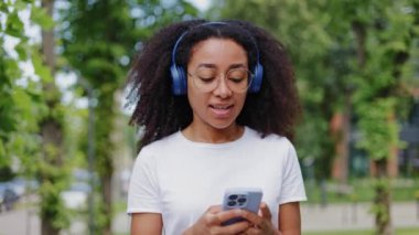 Gülümseyen Afro-Amerikan kadının ön görüntüsü açık havada yemyeşil çalıların yanında geziniyor ve kablosuz kulaklıklarla müzik dinliyor. Çekici genç bir kadın dışarıda akıllı telefonu kontrol ediyor..