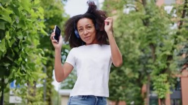 Mavi kablosuz kulaklıkla müzik dinleyen ve sokaklarda yeşillikle dans eden neşeli genç bir kadın. Canlı Afrikalı Amerikalı kadın modern akıllı telefonu elinde tutuyor ve dışarı doğru enerjik bir şekilde hareket ediyor..