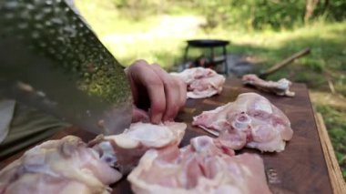 Bir şefin elleri ahşap bir kesme tahtasının üzerinde ustalıkla taze, çiğ tavuk hazırlıyor. Sahne yemek pişirme, yemek hazırlama ve açık havada yaşama temalarını çağrıştırıyor..