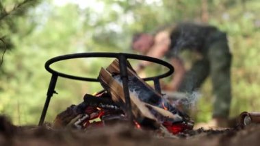 Kamp ateşinde yanan odunların yakın çekimi ve arka planda onunla ilgilenen bir adam.