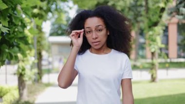 Çekici, aktif, Afro-Amerikan kadın ağaçların yanında yürüyor ve açık havada camları ayarlıyor. Sıradan beyaz tişörtler giymiş neşeli genç bir kadın kameraya bakıyor..