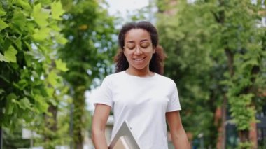 Beyaz tişört giymiş neşeli Afro-Amerikan bayan öğrenci Central Park 'taki yeşil ağaçların arasından geçiyor. Elinde dijital teknoloji ve günlük tutan olumlu bir kadın. İnsan kavramı.