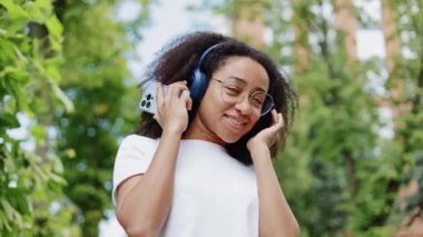 Mavi kablosuz kulaklıkla müzik dinleyen ve yeşil sokakta dans eden neşeli genç bir bayan. Aktif Afro-Amerikan kadın modern akıllı telefonu elinde tutuyor ve dışarı doğru enerjik bir şekilde hareket ediyor..