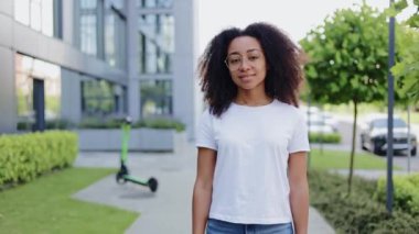 Temiz gözlükler takan neşeli Afro-Amerikan kadın kameraya modern binayla ve arka planda elektrikli scooterla bakıyor. Dişleriyle gülümseyen güzel bir kadın. Portre kavramı.