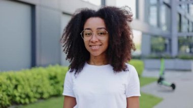 Çimen ve çalılarla modern binanın avlusunda dikilirken kameraya bakan pozitif Latin kadın. Yuvarlak gözlü mutlu mülk sahibi iş yeri kompleksi alanında yürüyor..