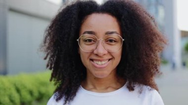Yakın plan ve modern binanın arka planında kameraya bakan temiz gözlüklü mutlu Afro-Amerikan kadın portresi. Çekici esmer kadın dişleriyle hafifçe gülümsüyor..