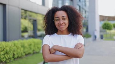 Pozitif ve çekici Afro-Amerikan kadının portresi. Kollarını kavuşturup kameraya bakıyor. Hoş bir gülümsemeyle. Kıvırcık kahverengi saçlı ve açık havada giyinmiş güzel bir bayan..