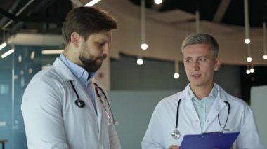 İki erkek doktor modern bir hastanede hastaların tıbbi çizelgelerini ve tedavi planlarını tartışıyor..