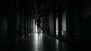 Karanlık bir ofis koridorunda tek başına yürüyen bir iş adamının silüeti. Parlak bir pencereye doğru, binadan çıkarken..