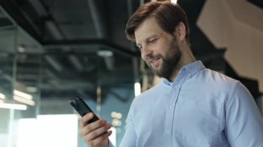 Modern ofiste çalışırken telefonuna mesaj atan mutlu iş adamı. Mali danışman broker yöneticisi mesaj atıyor, akıllı telefonla konuşuyor, ofis penceresinin önünde gülümsüyor, olumlu duygular besliyor.