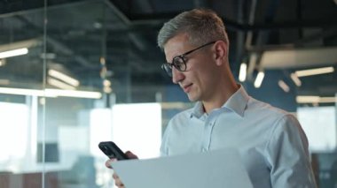 İş adamı telefonuna bakıyor ve elindeki belgeleri inceliyor. Gözlüklü erkek SEO uzmanı ofis odasında cep telefonu tutarken dokümanlara dayalı bir rapor hazırlıyor..