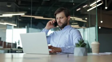 Meşgul işadamı, modern bir ofiste masasında oturuyor, dizüstü bilgisayarıyla çalışıyor ve telefonuyla konuşuyor. İşadamı erkek işçi odaklanmış ve üretkendir..