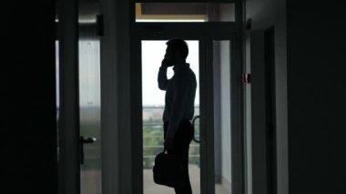 Bir iş adamının silueti telefonda konuşuyor, bir ofis binasının penceresinde duruyor..