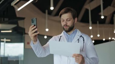 Bir erkek doktor elinde hasta dosyası tutarak internetten danışmanlık yapıyor ve bir video görüşmesinde konuşuyor. Tıbbi kayıtları uzaktan tartışıyorlar..